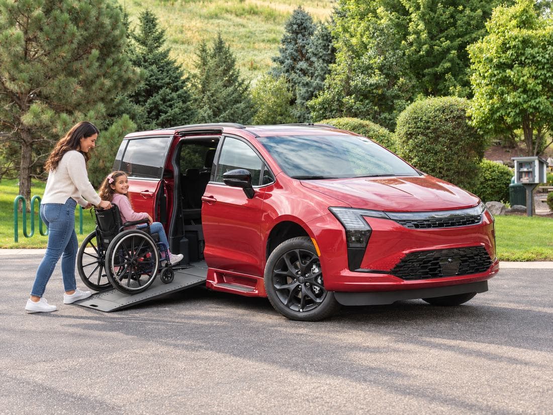 A mother and her young daughter, who uses a wheelchair, at the deployed side-entry ramp of a red Chrysler Pacifica on a neighbourhood street
